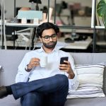 Indian business man using mobile phone reading news drinking coffee in office.