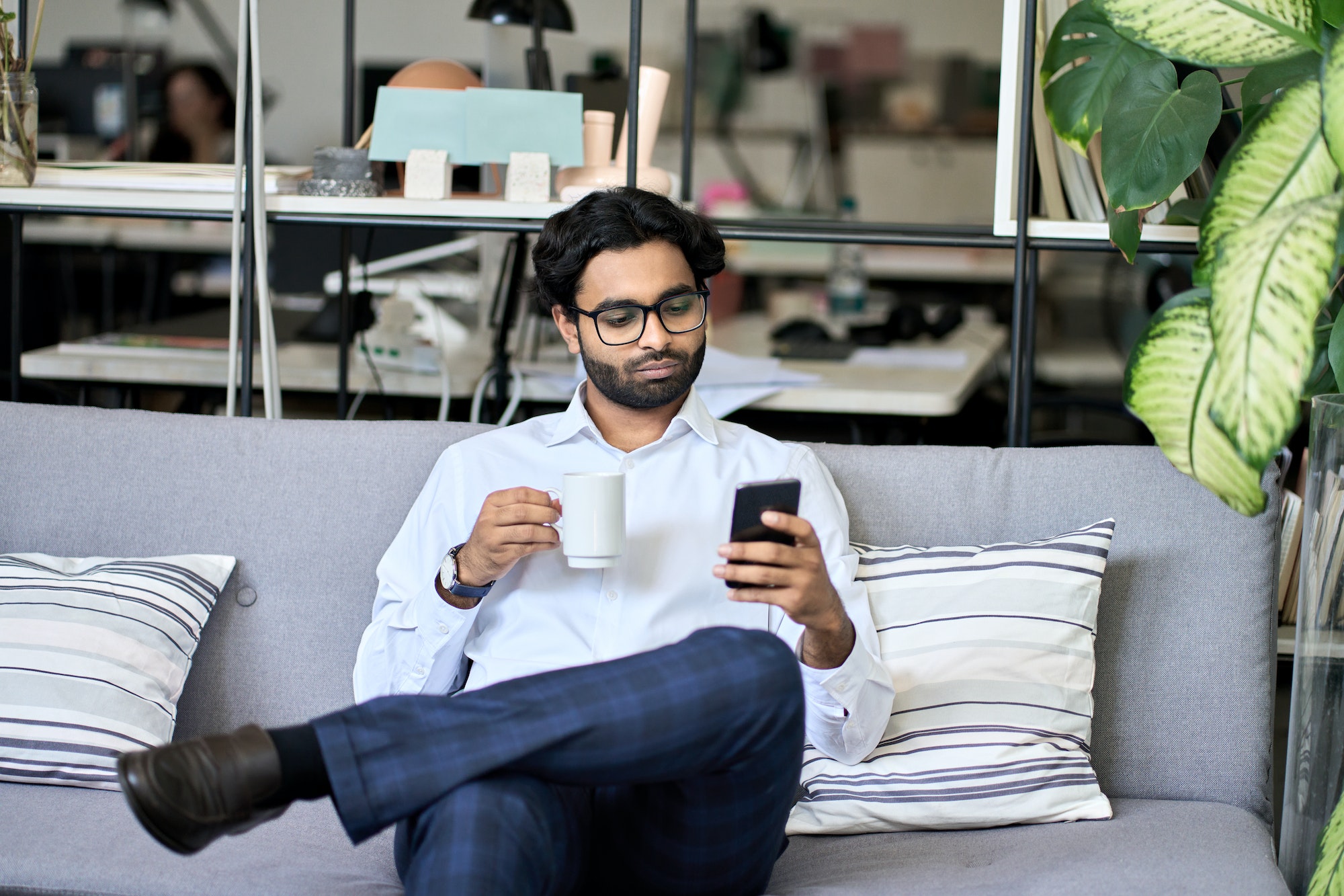 Indian business man using mobile phone reading news drinking coffee in office.